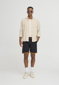 Lys beigem i skjorte med knapper, hvit t-skjorte, marineblå shorts med brune detaljer, og hvite sneakers. Tilbehør inkluderer solbriller og et brunt belte.