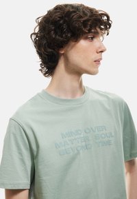 Camiseta de algodón verde claro con cuello redondo y un logo de texto texturizado en un tono ligeramente más oscuro, ubicado de forma centrada en la parte frontal.