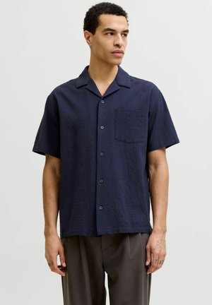 Muž v námorníckom modrom košeli s krátkym rukávom na gombíky a tmavých nohaviciach, stojaci pred jednoduchým svetlým pozadím, pozerajúci mierne doprava.