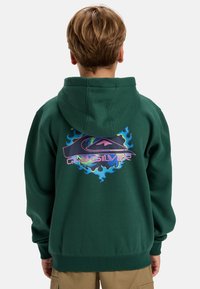 Sudadera verde con un gran gráfico multicolor que presenta texto y llamas en la parte posterior. Tejido suave, bolsillo canguro delantero y capucha con cordón.