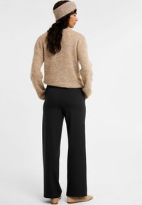 Beige fuzzy trui met een losse pasvorm, gecombineerd met zwarte wijde broek en beige instappers, gedragen met een bijpassende hoofdband.