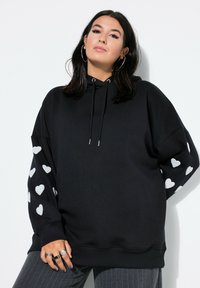 Sudadera negra con un ajuste oversize, que presenta patrones de corazones blancos en las mangas, capucha con cordón y dobladillo acanalado. Se destaca su textura de tela suave.