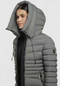 Giacca imbottita grigia con un cappuccio grande e strutturato, chiusura con zip davanti e tasche laterali. Presenta sezioni trapuntate pronunciate e dettagli con cordoncino.