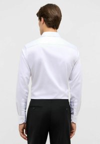 Mann mit dem Rücken zur Kamera, der ein langärmliges weißes Hemd trägt, das in schwarze formelle Hosen gesteckt ist, vor einem schlichten Hintergrund.