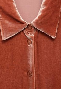 Camicia di velluto color ruggine con colletto, impreziosita da due bottoni marroni sul davanti e una consistenza morbida e liscia.