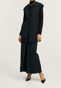 Chaleco largo azul marino con corte entallado, diseño sin mangas y bolsillos laterales, combinado con pantalones de pierna ancha y tacones negros puntiagudos.