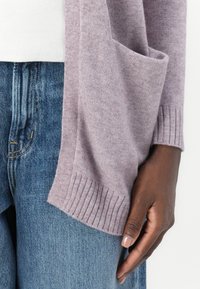 Hellblau-lavendelfarbener, weicher Strickcardigan mit einer Fronttasche und gerippten Bündchen, getragen über einem weißen Oberteil und blauen Jeans. Die Textur wirkt glatt.