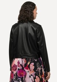 Giacca nera in finta pelle con un design aderente, caratterizzata da spalline e una vita con cinturino, indossata su un vestito a fantasia floreale.