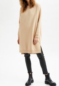 Beige oversized sweatshirtklänning med långa ärmar och sidofåror, i kombination med svarta slim-fit byxor och kraftiga svarta ankelboots.