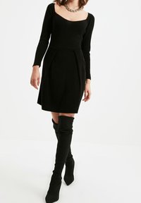 Black dress with a square neckline, long sleeves, and a pleated skirt. Paired with knee-high black boots and a chunky chain necklace.