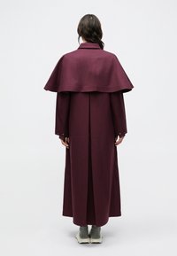 Maroon long coat with a cape-style overlay, tailored collar, long sleeves, and buttoned cuffs. Smooth texture, minimalistic design.