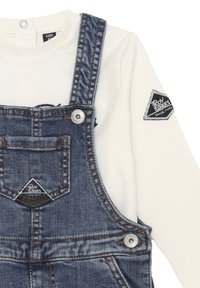 Salopette in denim blu medio, con tasca frontale, accessori in argento e una maglietta a maniche lunghe bianca con una toppa nera con logo sulla manica.