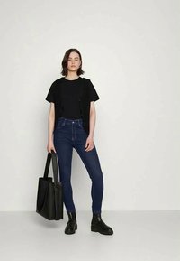 Cardigã preto canelado sobre uma t-shirt preta, combinado com jeans skinny azul escuro e botas pretas de tornozelo, segurando uma bolsa tote preta.