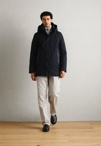 Chaqueta de plumas de color azul marino oscuro con capucha, que cuenta con dos bolsillos laterales con cremallera. Combinada con pantalones gris claro y zapatos negros, de pie sobre un suelo de madera.