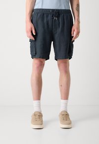 Shorts en lin bleu foncé avec une taille élastique, un cordon de serrage et des poches latérales. Portés avec des baskets beiges et des chaussettes blanches montantes.
