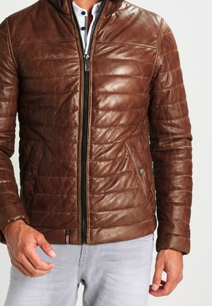 Homme portant une veste en cuir matelassée marron zippée sur une chemise henley blanche et un pantalon gris clair.