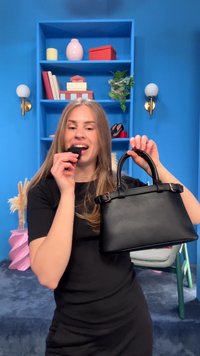 Jeune femme tenant un sac à main noir dans une main et un petit objet noir près de sa bouche, debout dans une pièce colorée avec des étagères.