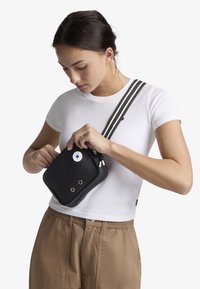 Borsa a tracolla nera con una texture liscia, cinturino regolabile a righe, chiusura con zip e un'etichetta rotonda con logo sulla parte anteriore.