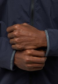 Marineblau leichte Jacke mit Reißverschluss, versehen mit grauen Besätzen an den Bündchen. Nahaufnahme von zwei Händen, die den Saum der Jacke halten.