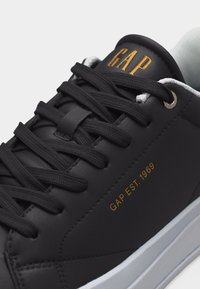 Baskets en cuir noir avec lacets noirs, semelle blanche, texte doré "GAP EST. 1969" sur le côté et "GAP" sur la languette.