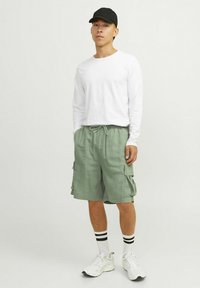 Chemise blanche à manches longues, short cargo vert clair avec poches latérales, casquette de baseball noire, chaussettes rayées noires et blanches, et baskets blanches.