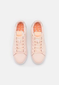 Un paio di sneakers lace-up rosa chiaro con suole bianche e plantari arancioni che presentano testo bianco su uno sfondo bianco.