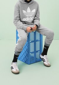 Šedá mikina s bílým logem Adidas, odpovídající tepláky s bílými pruhy, černé ponožky s logy a bílé tenisky s hnědými detaily. Sedí na modré bedně.