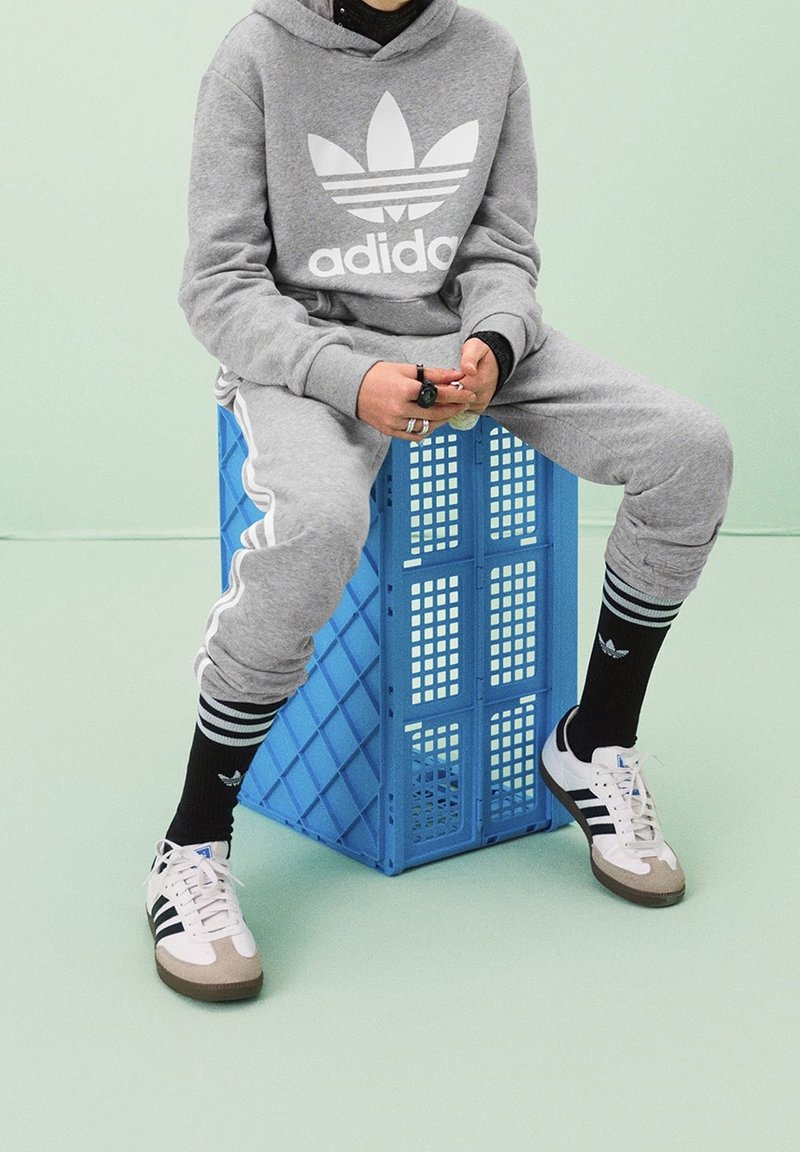 Šedá mikina s bílým logem Adidas, odpovídající tepláky s bílými pruhy, černé ponožky s logy a bílé tenisky s hnědými detaily. Sedí na modré bedně.