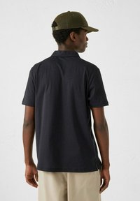 Polo nero con maniche corte, texture liscia e colletto classico. Indossato con pantaloni khaki e un cappello verde oliva.