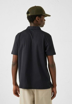Zwart polo shirt met korte mouwen, gladde textuur en een klassieke kraag. Gekozen in combinatie met kaki broeken en een olijfgroene pet.