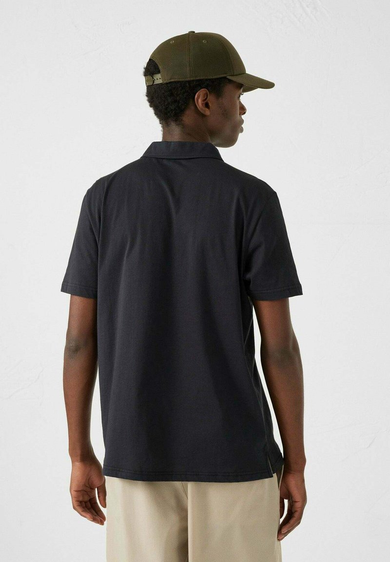 Polo nero con maniche corte, texture liscia e colletto classico. Indossato con pantaloni khaki e un cappello verde oliva.