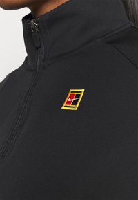 Chaqueta negra con cremallera y cuello alto, con un logo bordado multicolor en el lado izquierdo. Tela suave y ajustada con costuras limpias.
