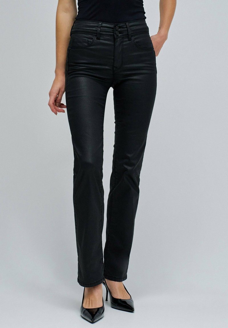 Kobieta w dopasowanych czarnych spodniach z wysokim stanem i czarnych czółenkach na wysokim obcasie, stoi z jedną ręką w kieszeni na tle jednolitego tła.