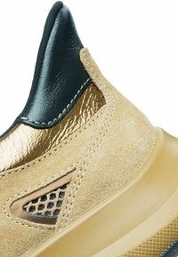 Sneaker in suede color tan con una parte superiore in pelle nera, accenti in oro metallico, ritagli in rete e una suola in gomma chiara con grip testurizzato.
