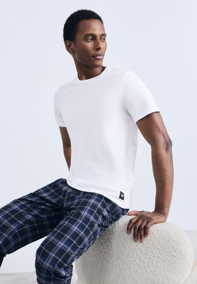 Balta kokvilnas T-krekls ar īsām piedurknēm; kopā ar tumši ziliem un zilā pārbaudāmajiem biksēm. Modelis sēž uz teksturēta, bēša skulpturāla ottomana.