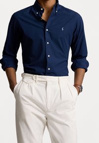 Navyblaues Hemd mit Knopfleiste, das einen klassischen Kragen und ein hellblaues Logo aufweist, kombiniert mit weißen, plissierten Hosen und einer silbernen Uhr.