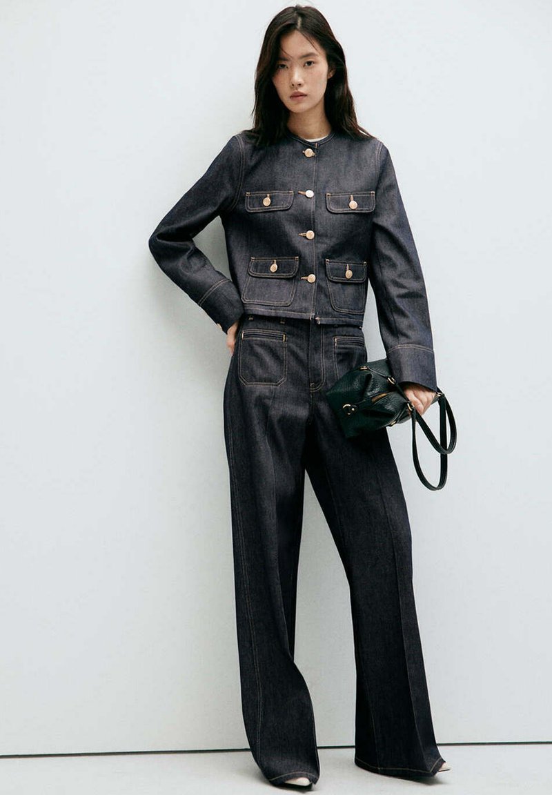 Femme portant une veste en jean foncé avec des boutons dorés et un pantalon large assorti, tenant un petit sac noir à ses côtés.