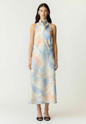 Femme portant une robe sans manches, col montant, longueur cheville avec un motif floral bleu pastel, pêche et crème, associée à des talons noirs ouverts à l’avant.