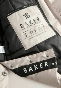 Etichetta interna di una giacca trapuntata nera e crema che presenta il nome del marchio "BAKER by TED BAKER", con istruzioni per la cura e simboli per la resistenza all'acqua e il calore.