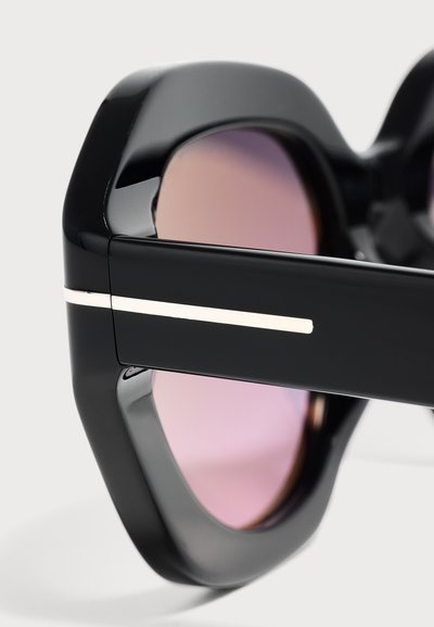 Gros plan de lunettes de soleil noires avec des verres teintés roses et une fine bande blanche sur les branches épaisses sur un fond neutre.