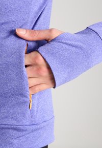 Maglietta sportiva viola aderente con un aspetto testurizzato, dotata di una tasca con cerniera sul lato sinistro e fori per il pollice nei polsini.