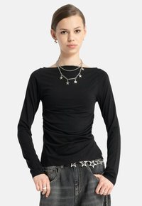 Młoda kobieta w czarnej bluzce z długim rękawem i szarych dżinsach, nosząca warstwowe srebrne naszyjniki w kształcie gwiazdy oraz pas z łańcuchem w tym samym stylu, z rękami w kieszeniach.