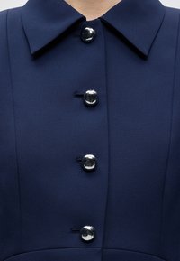Vêtement bleu foncé boutonné avec quatre boutons ronds argentés brillants et un large col, présenté sur le haut du torse d’une personne.