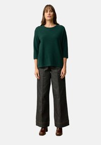 Maglione verde a maglia con maniche tre quarti, abbinato a jeans neri a gamba larga con cuciture gialle. Stivali con tacco marroni completano il look.