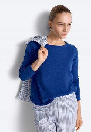 Žena s vlasmi stiahnutými dozadu nosí modré tričko s dlhým rukávom a pruhované nohavice, drží pruhovanú košeľu cez rameno a pozerá sa do strany.