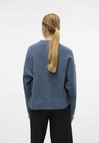 Kvinna med långt blont hästsvans som bär stora hoopsmycken, en blå tröja och svarta byxor, stående vänd mot en enkel vit bakgrund.