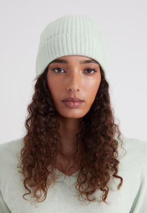 Junge Frau mit langen, lockigen braunen Haaren, die eine hellgrüne Strickmütze und einen passenden Pullover trägt, blickt geradeaus mit einem neutralen Ausdruck.