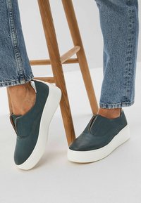 Blaue Leder-Slipper mit weißen Gummisohlen, die an der Oberseite einen goldenen Akzent und eine glatte Textur aufweisen, kombiniert mit Denim.