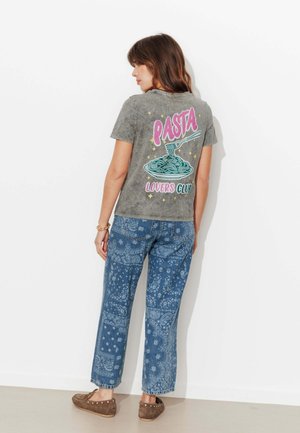 Femme portant un t-shirt gris avec l'inscription "Pasta Lovers Club" et un motif de fourchette enroulant des spaghettis, accompagné d'un jean bleu à motifs et de chaussures marron.