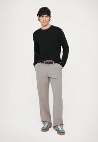 Camisola preta, calças cinza-claro, cinto castanho entrançado e ténis verdes. Modelo em pé, com as mãos nos bolsos, fundo neutro.